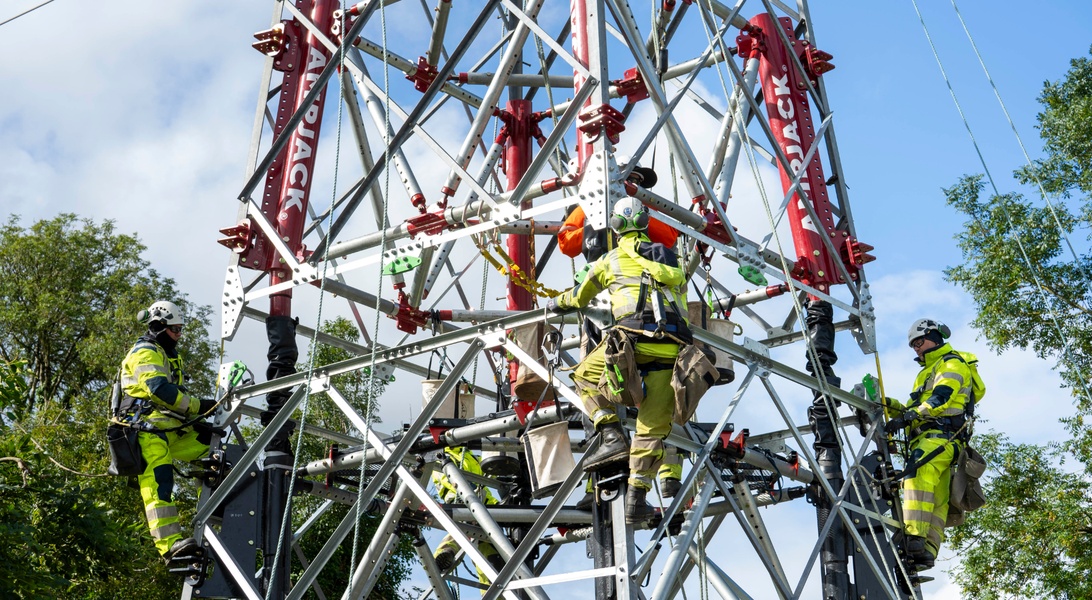 Utility workers assembling a high-voltage electricity transmission tower surrounded by steel framework and trees under a blue sky.