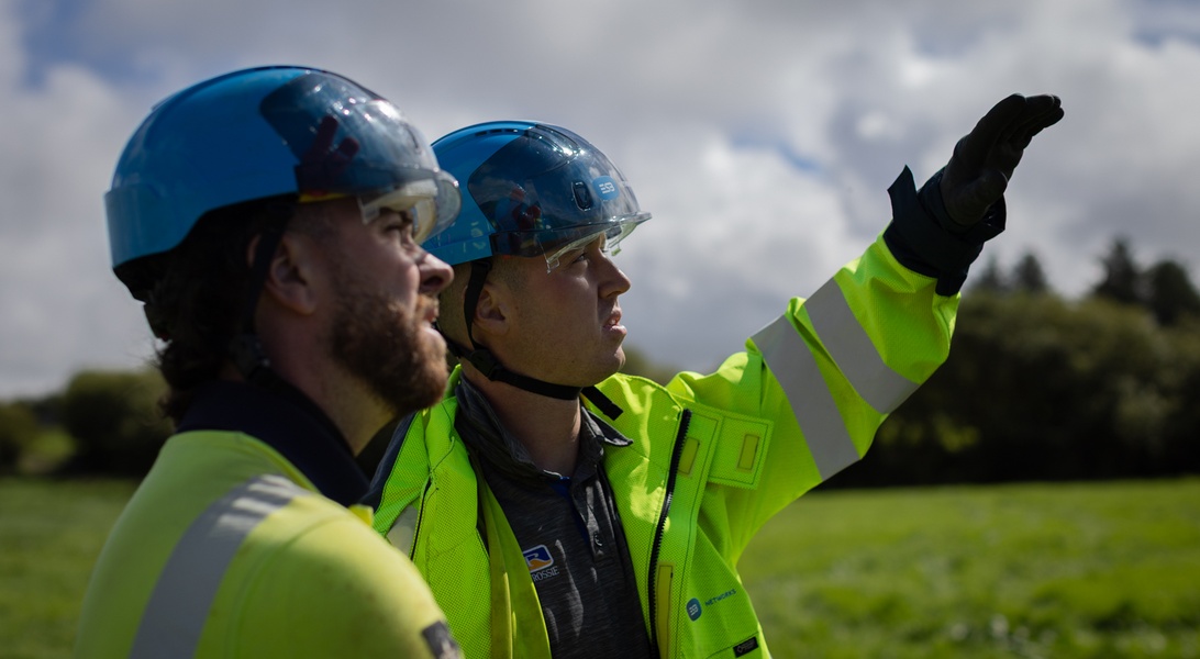 Two network technicians in high-visibility jackets and blue safety helmets standing outdoors in a grassy field. One worker is pointing toward the horizon while the other looks in the same direction.