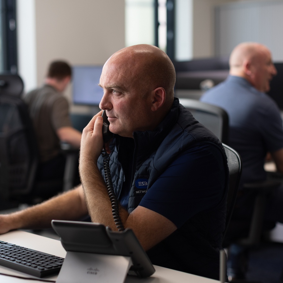 ESB Networks employee working in an office environment, speaking on a desk phone at a computer workstation, with colleagues working nearby.