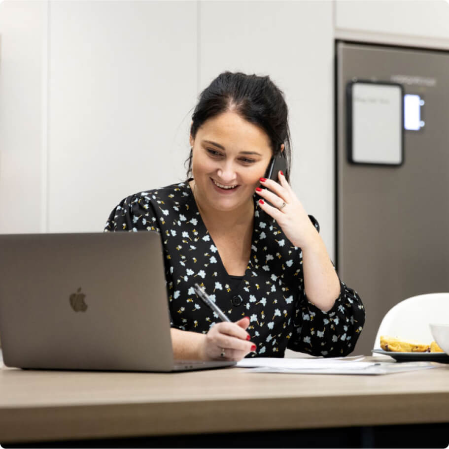 A woman seated at a table, holding a phone with a laptop beside her